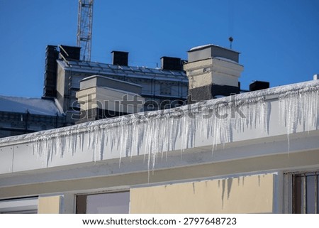 Rows of sharp icicles hang from snow-covered eave of urban residential building with chimneys and construction crane visible against clear blue winter sky.
