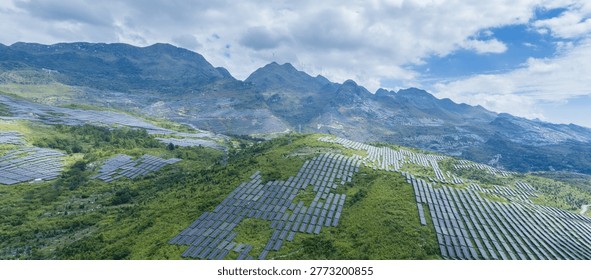 Rows of solar panels blanket rolling green hills under a partly cloudy sky; distant wind turbines crown ridges in this large-scale renewable energy site. 