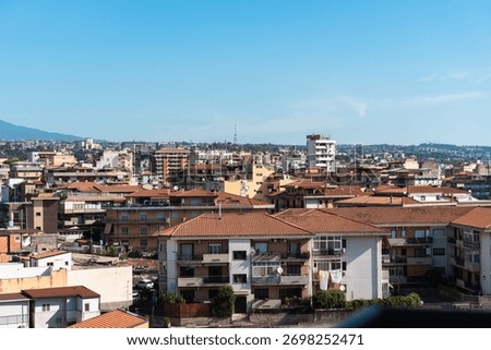 Rows of terracotta rooftops stretch across Catania with Mount Etna visible on the horizon. The contrast of warm buildings and cool blue sky reflects the vibrant Sicilian atmosphere.