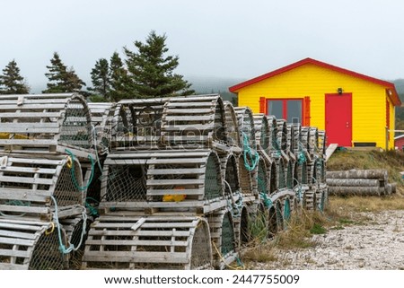 Rows of traditional wooden lobster pots and traps stacked on a grassy meadow. The cages are made of small wood sticks, green nylon fishing rope. There's a yellow storage building with a red door.  