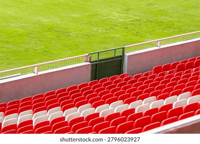 Rows of vibrant red and white seating fill the tiered stand of an empty stadium, positioned above a bright green sports field and a protective railing under the daytime sun
