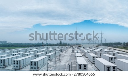 Rows of white battery container units line paved aisles at grid-connected energy storage facility under construction, with transmission towers and substation infrastructure in the background