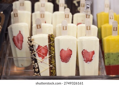 Rows of white cream popsicles featuring embedded strawberry slices and chocolate nut toppings in a refrigerated display case.