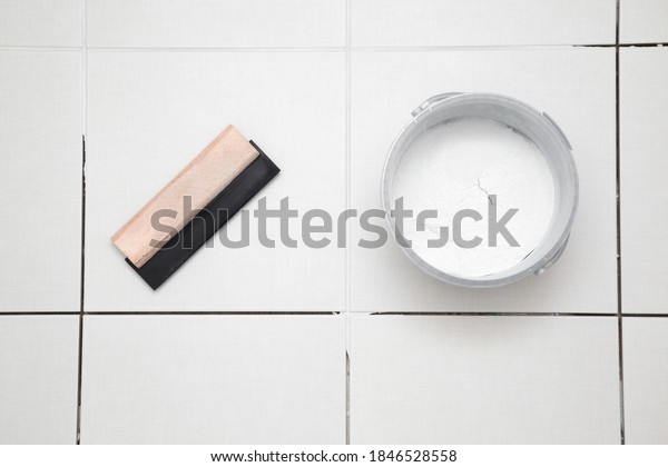 Rubber trowel and container\
with powder of grouting paste for ceramic tile seams on floor.\
Closeup. 