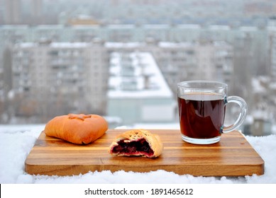 Ruddy bitten pie with filling, a glass mug of hot black coffee, and a wooden cutting board sit on a snow-covered roof parapet against a blurred background of a winter morning city.