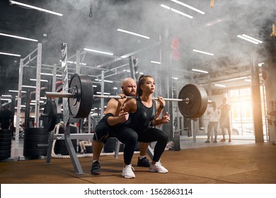 Rude brutal trainer with bald head and thick brunette beard standing behind young female powerlifter, backing her up, controlling process of squatting using heavy barbell in gym, white smoke in air