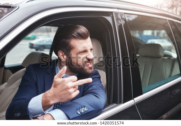 Rude businessman. Angry and rude bearded businessman\
in formal wear is gesturing and making a grimace while driving his\
car.