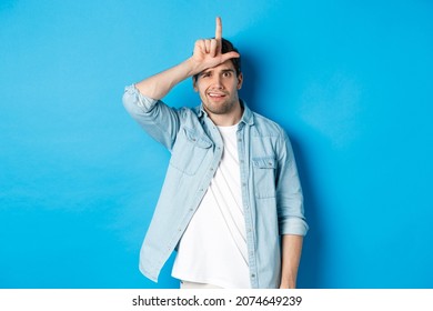 Rude Guy Mocking Person For Losing, Showing Loser Sign On Forehead And Looking With Dismay, Standing Against Blue Background