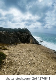 Rugged cliffs and a distant lighthouse overlook the Atlantic under stormy skies near Portugal.