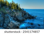 A rugged cliffside in Acadia National Park, Maine, overlooks the deep blue ocean. The rocky shore, dotted with evergreens, creates a stunning contrast against the calm coastal waters.