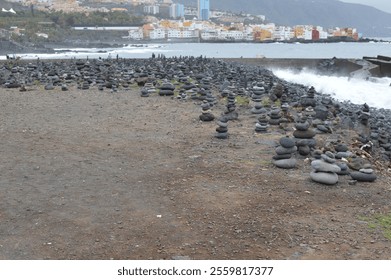 A rugged coastal landscape featuring numerous carefully balanced stone stacks on a rocky beach. Waves crash against the shore in the background, with colorful coastal buildings and hills - Powered by Shutterstock