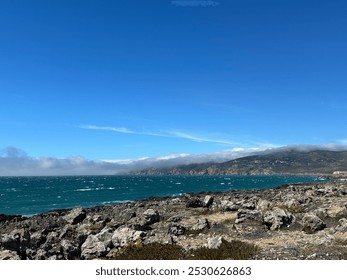 A rugged coastal landscape with rocky shores and a deep blue ocean stretching into the horizon. The mountains in the distance are partly covered by misty clouds under a clear blue sky, serene seascape - Powered by Shutterstock