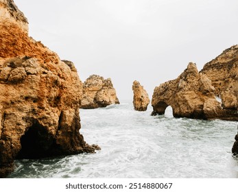 Rugged coastal rock formations rising from the ocean, with natural arches and cliffs shaped by the waves, creating a striking seascape against the backdrop of a cloudy sky - Powered by Shutterstock