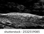 Rugged landscape with jagged, rocky terrain under a dramatic, cloud-filled sky, creating a stark contrast in black and white tones.