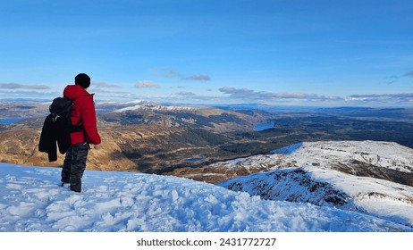 A rugged male hiker, clad in winter attire, stands atop a snow-dusted mountain peak, gazing at the majestic landscape. The tranquil lake, clear sky, and distant mountain ranges. Travel adventure alone - Powered by Shutterstock