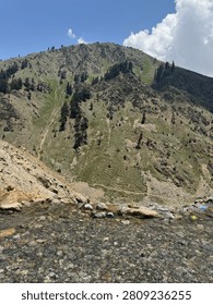 A rugged mountain landscape with sparse trees and a rocky foreground under a blue sky.
