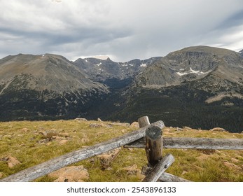 A rugged mountain landscape stretches under a cloudy sky, with rocky peaks partially covered by patches of snow. A rustic wooden fence in the foreground adds depth to the expansive view. - Powered by Shutterstock