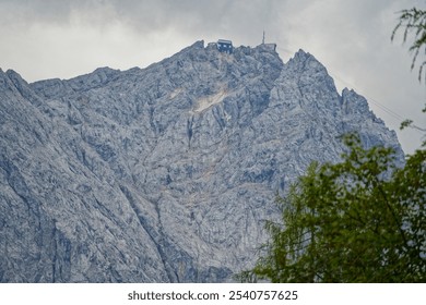 A rugged mountain peak with a small building at the summit, surrounded by rocky terrain and cloudy skies. Lush greenery is visible in the foreground. - Powered by Shutterstock