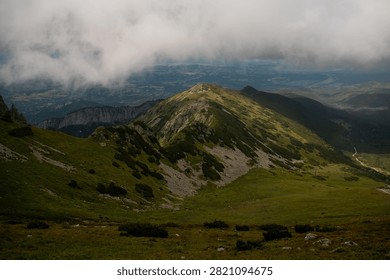 A rugged mountain range with a sharp, rocky crest is partially obscured by low-hanging clouds, revealing a vast valley landscape