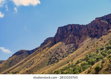 Rugged mountain range under clear blue sky. Slopes covered in dry grass and shrubs. Bright sunlight highlights rocky ridges