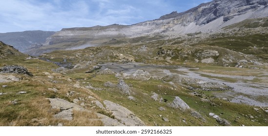A rugged mountain valley with rocky terrain and a small stream, under a cloudy sky.
