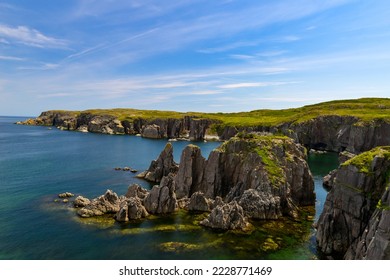 A rugged ocean coastline with high rocky sea stacks, cliffs, vibrant green grass, blue sky, and clouds. The smooth water is a blue color. There's a hiking trail along the edge of the cliff and coast.  - Powered by Shutterstock