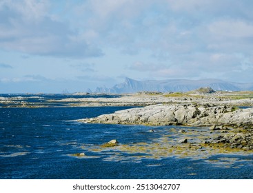 A rugged rocky coastline on Andøya, Northern Norway, with distant mountains under a cloudy sky and waves crashing along the shore - Powered by Shutterstock