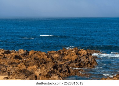 A rugged rocky shoreline meets the deep blue ocean under a clear sky, with gentle waves breaking against the rocks - Powered by Shutterstock