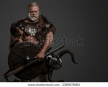 A rugged, silver-haired, bearded elderly viking dressed in fur and lightweight armor, with a helmet attached to his belt, holding an axe and shield against a gray background