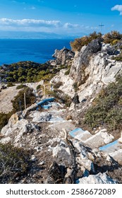 A rugged stone path with blue-painted steps leads down a rocky hillside, overlooking the Corinthian Gulf and Melagavi Lighthouse, with a cross on the cliff.