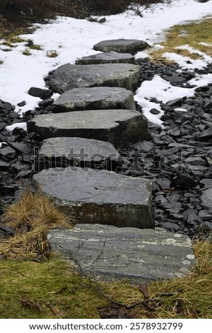 A rugged stone pathway winds through a natural setting, partially covered by melting snow, revealing dark rocks beneath. 