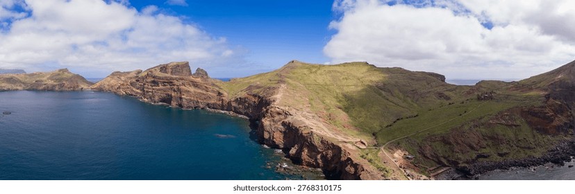 Rugged volcanic cliffs drop into the Atlantic at Ponta de Sao Lourenco, Madeira. A narrow hiking path traces ridgelines toward jagged sea stacks under midday sun.