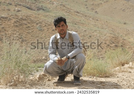 Ruggedly handsome Asian model Zainullah setting proudly on a mountaintop, wearing a flowing shalwar qameez, black shoes, and carrying a gray bag.
