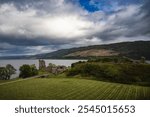 Ruined Urquhart Castle in the Highlands of Scotland besides Loch Ness lake in background with dramatic sky and part of rainbow.