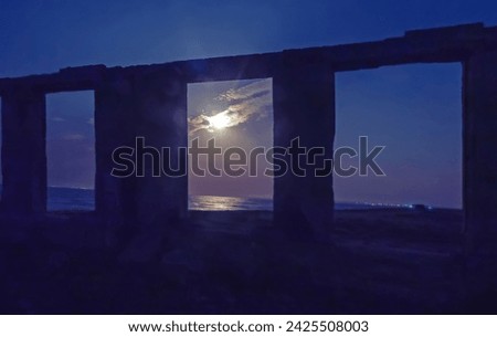 Ruins by the sea in the moonlight. The full moon is shining through an window opening. Lights in the far coastal village are visible