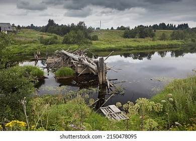 Ruins Wooden Abandoned Road Bridge Across The River Against The Backdrop Of The Summer Landscape And Overcast Sky. The Construction Of Logs Is Destroyed. Grass Grass, Tree Branches Brought By Flow Of 