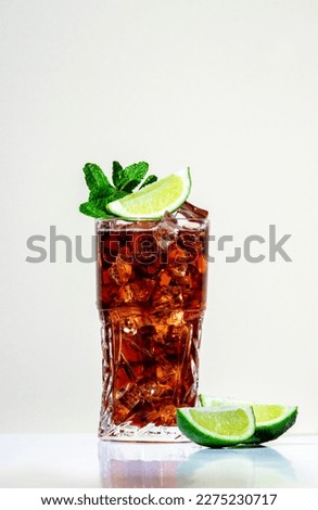 Rum cola cocktail with strong alcohol and ice, garnished with mint and lime in highball glass. Beige background, hard light