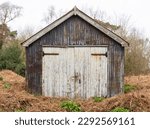 Run down garage building 
shed made of rusty corrugated panels, with old white wooden doors, surrounded with overgrown vegetation. Suffolk. UK