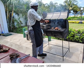 Runaway Bay, Saint Anns, Jamaica. March 21, 2019. A chef cooks Jerk Chicken on a traditional oil drum barbeque grill in front of palm trees, a lawn and  sea. 