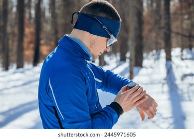 Runner Checking Smartwatch During Winter Training in Snowy Forest. A runner man in a blue jacket and headband pauses to check his smartwatch during a winter training session in a snowy forest setting.