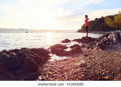 Runner Resting From Routine Exercise. Happy Jogger Relaxing At The Beach After Run, Standing On The Stones And Looking At The Sea. Active Young Woman Enjoying Sunrise.