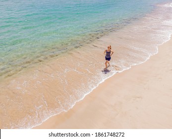 Runner Running On Beach By The Ocean - View From Above. Woman Athlete Training Cardio Jogging Doing Morning Workout. Hero Aerial Drone View Shot, Lots Of Copy Space