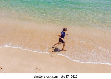 Runner Running On Beach By The Ocean - View From Above. Man Athlete Training Cardio Jogging Doing Morning Workout. Hero Aerial Drone View Shot, Lots Of Copy Space