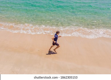 Runner Running On Beach By The Ocean - View From Above. Man Athlete Training Cardio Jogging Doing Morning Workout. Hero Aerial Drone View Shot, Lots Of Copy Space