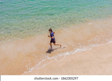 Runner Running On Beach By The Ocean - View From Above. Man Athlete Training Cardio Jogging Doing Morning Workout. Hero Aerial Drone View Shot, Lots Of Copy Space