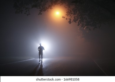 Runner Running On A Dark Misty Night Through Tree Lined Streets, Silhouetted By Street Lights. 