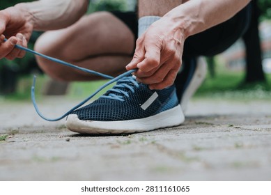 A runner ties the laces of a blue athletic sneaker while crouched on a paved path. Close-up view shows hands fastening the shoe before an outdoor run.