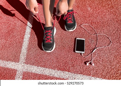 Runner Woman Athlete Tying Running Shoes Laces Getting Ready For Race Training On Run Track With Smartphone And Earphones For Music Listening On Mobile Phone.