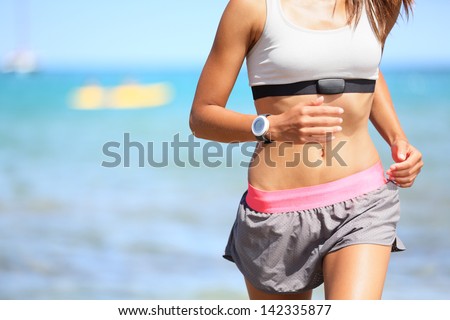 Runner woman with heart rate monitor running on beach with watch and sports bra top. Beautiful fit female fitness model training and working out outside in summer at part of healthy lifestyle.