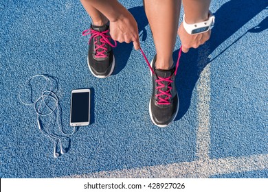 Runner woman tying running shoes laces getting ready for race on run track with smartphone and earphones for music listening on mobile phone. Athlete preparing for cardio training. Feet on ground. - Powered by Shutterstock